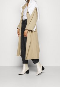 Beżowy długi płaszcz o szerokich rękawach, noszony na białej koszuli, zestawiony z ciemnymi dżinsami i błyszczącymi białymi botkami na kostkę z blokowym obcasem.