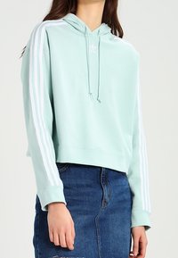 Vaalean mintunvihreä lyhyt huppari, jossa on valkoiset kolmoisraidat hihoissa, kiristysnauhallinen huppu ja pieni logo rinnassa. Alta näkyy farkkuhame.