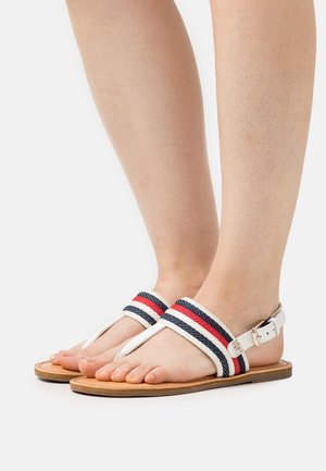 Des pieds portant des sandales blanches avec des sangles en tissu rayé rouge, bleu et blanc et une semelle marron, présentés sur un fond blanc uni.