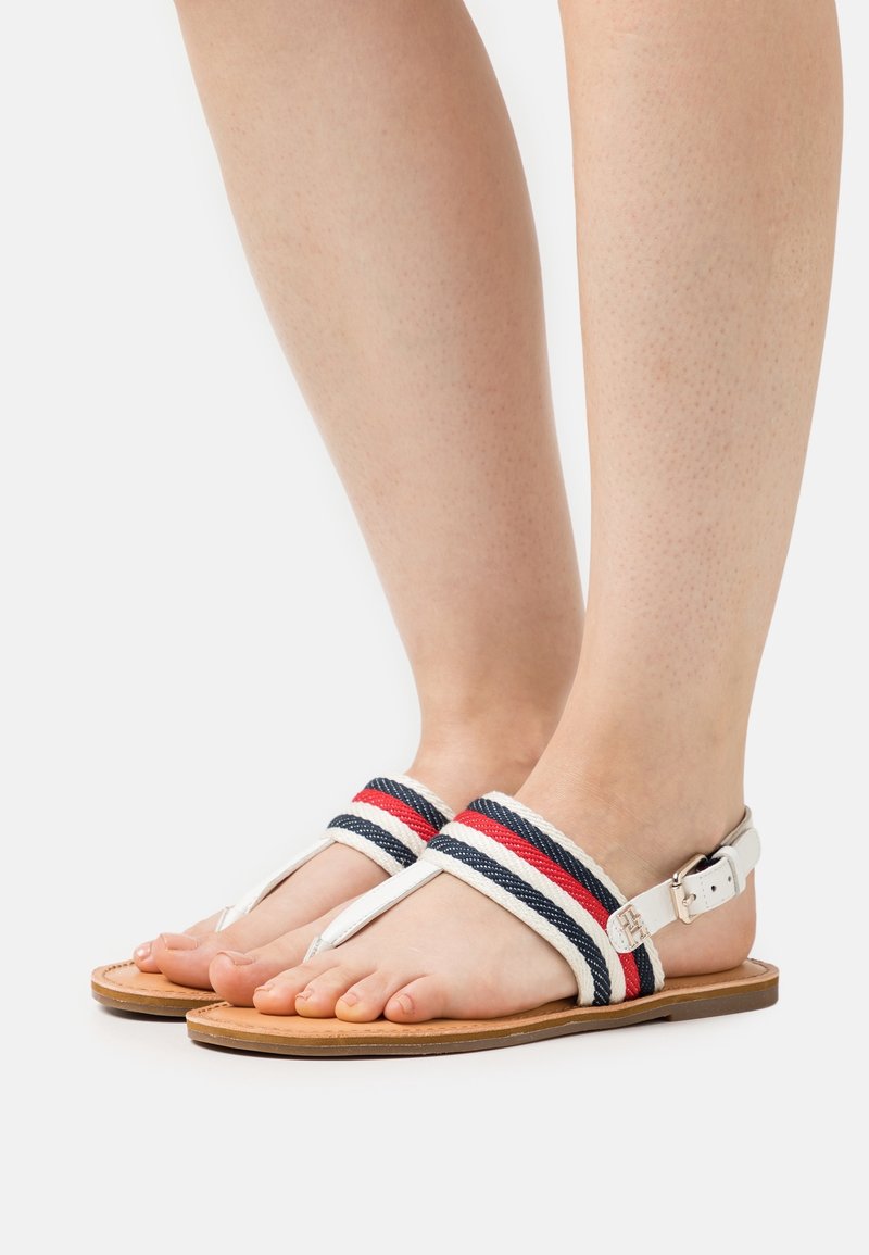 Piedi che indossano sandali bianchi con cinturini in tessuto a righe rosse, blu e bianche e una suola marrone, mostrati su uno sfondo bianco semplice.
