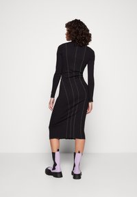 Schwarzes knielanges Kleid mit langen Ärmeln, verziert mit grüner Stickerei an den Seiten; kombiniert mit lila und schwarzen Knöchelstiefeln.