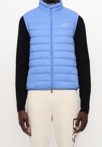 Světle modrá prošívaná vesta s vysokým límcem, předním zipem a logem na levé hrudi. Nositá přes černý top s dlouhým rukávem.