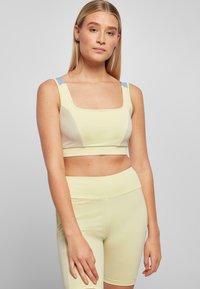 Žuti sportski crop top s kvadratnim izrezom i svijetloplavim akcentima na ramenima, u kombinaciji s visokim žutim šorcama. Gladak materijal, usklađen dizajn.