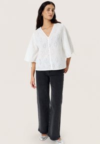 Hvid broderet bluse med V-udskæring, diamant-formede udskæringer og brede ærmer, parret med højtaljede sorte jeans og sølvfarvede flade sko.