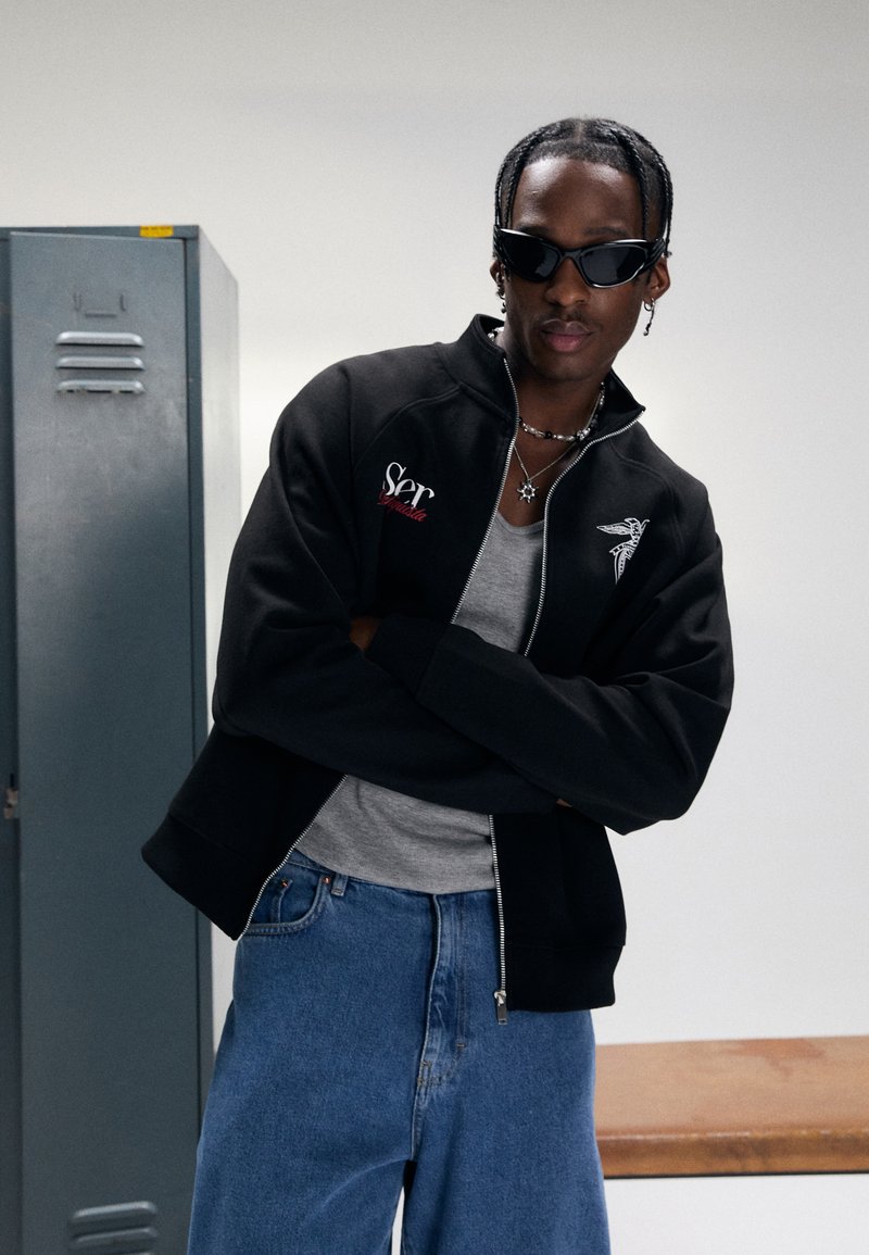Young man with sunglasses, braided hair, black zip-up jacket, gray shirt, and blue jeans standing with arms crossed near a locker and bench.
