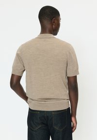 Ein Mann, der nach hinten schaut und ein beiges, kurzärmeliges Strickshirt und dunkle Jeans aus Denim vor einem schlichten weißen Hintergrund trägt.