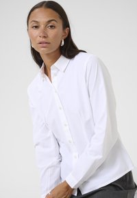 Chemise blanche à col pointu, à manches longues et à la texture douce. Avec un système de fermeture à boutons et une coupe décontractée avec des fentes latérales.