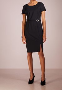 Femme portant une robe noire ajustée à mi-genoux avec des manches courtes, une fente sur le côté, un détail de ceinture, des talons noirs et un sac à l'épaule.