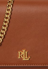 Brūna ādas rokassomiņa ar zelta ķēdes siksnu un zelta izgriezto "RLL" logotipu priekšpusē, ar gludu tekstūru un tīru dizainu.