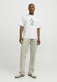 Tricou alb din bumbac, cu un imprimeu grafic verde și textul "Fără pete". Purtați cu blugi gri deschis, ușor modelați, și adidași bej.