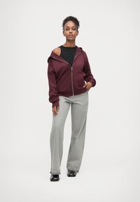 Femme debout, une main dans la poche, portant un sweat à capuche zippé bordeaux par-dessus un haut noir, un pantalon gris à jambes larges et des chaussures noires sur fond blanc.