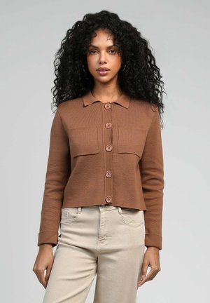 Femme aux cheveux bouclés portant un cardigan marron boutonné et un pantalon beige, debout devant un fond gris clair uni.