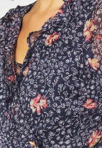 Blouse florale marine avec petites fleurs roses et bleues, col à volants et tissu léger et transparent pour une texture douce.