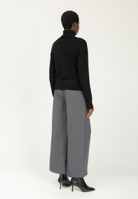 Schwarzer Rollkragenpullover mit geripptem Saum, kombiniert mit grauen weit geschnittenen Hosen. Das Model trägt schwarze Stiefeletten; glattes Material mit feiner Struktur.
