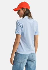 Hellblaues Baumwoll-Polohemd mit kurzen Ärmeln, präsentiert einen klassischen Kragen und Seitenschlitze, kombiniert mit hellblauen Jeans und einer orangenen Kappe.