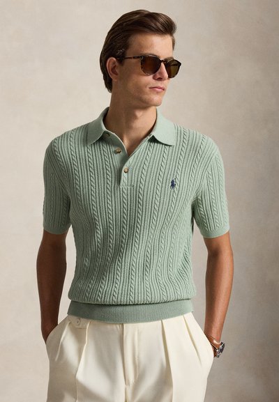Jeune homme portant des lunettes de soleil marron, polo en maille torsadée à manches courtes vert clair, pantalon plissé crème, mains dans les poches.