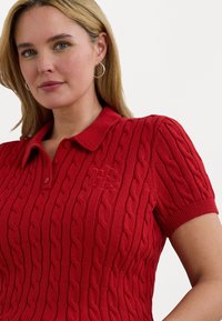 Rød strikket polo skjorte med korte ermer, med et strukturert kabelmønster og en krage. Brodert logo på venstre side.