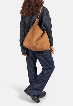 Sac à bandoulière en daim marron à forme courbe, avec un ornement décoratif en métal. La sangle est ajustable et en cuir.