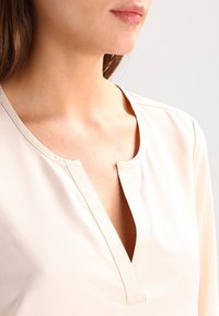 Top beige chiaro con scollatura a V, maniche lunghe e texture liscia. Presenta dettagli di cucitura sottili lungo il collo.