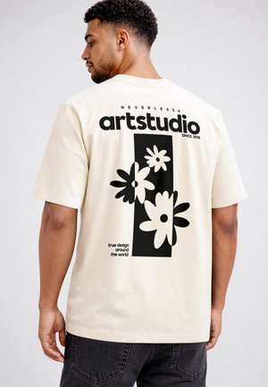 Man wearing a cream t-shirt with black floral graphic and text "neverless artstudio since 2016" and "true design around the world" on the back.