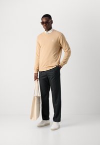 Suéter beige, pantalones oscuros, zapatillas blancas, sosteniendo un bolso crema. Con cuello redondeado, logo sutil en el pecho, textura suave.