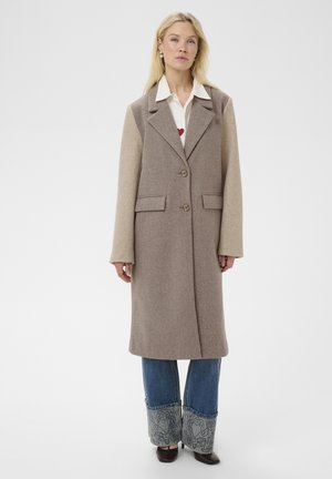 Femme portant un long manteau en laine beige bicolore sur un jean bleu avec des poignets brodés, debout devant un fond blanc uni.