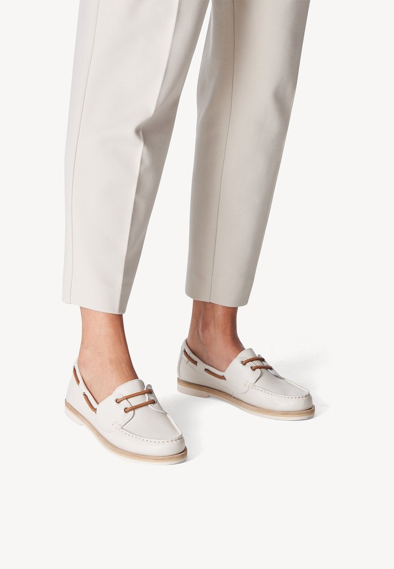 Scarpe casual bianche con finitura testurizzata, dotate di lacci e dettagli in pelle marrone, abbinate a pantaloni leggeri e alla caviglia.