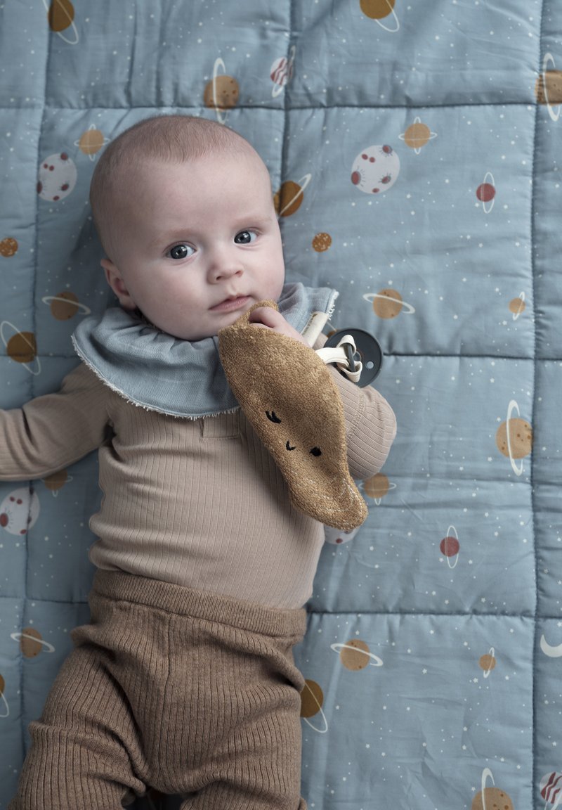 Mäkké, rebrované béžové body a hnedé nohavice; drží plyšovú hnedú hračku s usmievavou tvárou. Obklopený modrou prikrývkou s motívmi planét.