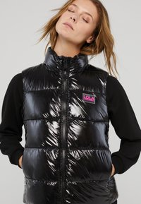 Musta puhvikuoriväri, jossa on kiiltävä pinta, korkea kaulus, etuvetoketju ja vaaleanpunainen "LOLA" -logo vasemmalla puolella.
