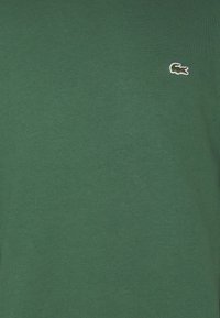 Maglietta in cotone verde con collo a giro, dotata di un piccolo logo di coccodrillo ricamato sul lato sinistro del petto. Texture liscia.