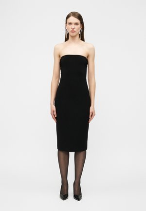 Femme portant une robe noire midi bustier avec des collants noirs transparents, des escarpins noirs pointus et de longues boucles d'oreilles pendantes en perles, se tenant devant un fond uni.
