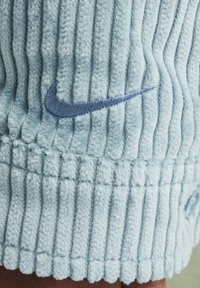 Šviesiai mėlynos velvetinės medžiagos, turinčios pakeltas vertikalias juosteles, kairėje pusėje yra tamsiai mėlynas siuvinėtas Nike logotipas. Minkšta tekstūra.