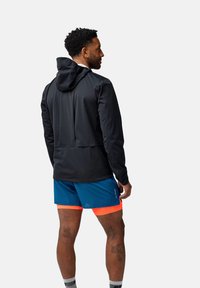 Črna kapuca z lahkim dizajnom, nosi se čez modre kratke hlače z oranžnimi poudarki. Gladka tkanina in preprost kroj.