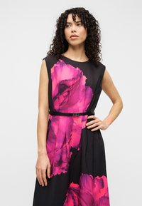 Robe noire sans manches ornée de grands motifs floraux roses, avec une taille cintrée et une fine ceinture noire mettant en valeur la silhouette.