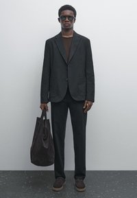 Veste noire cintrée, chemise marron foncé, pantalon noir et chaussures en daim marron. Tient un sac tote en daim marron. Stylisé avec des lunettes de soleil.