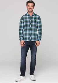 Camicia a quadri verde e blu con bottoni, abbinata a jeans in denim scuri e sneaker bianche. Include una toppa con logo sulla manica.