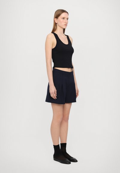 Schwarzes geripptes Tanktop kombiniert mit marineblauen, gefalteten Shorts. Das Model steht seitlich und präsentiert das figurbetonte Design und die knielangen Shorts.