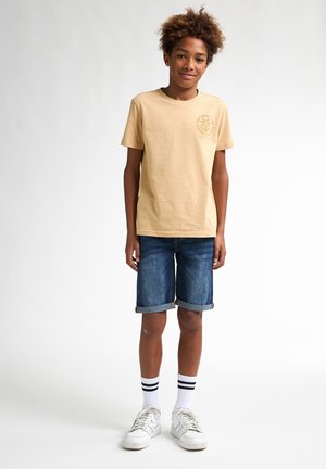 T-shirt in cotone beige con grafica di palme, abbinato a pantaloni shorts in denim blu con orli arrotolati. Sneakers bianche con strisce nere sui calzini.