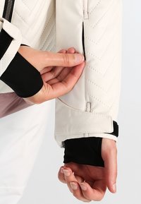 Manche de veste matelassée blanche à texture lisse, équipée d'une poche zippée et d'un poignet noir. Main montrant l'ouverture de la poche.