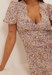 Kukkakuvioinen mekko, jossa on syvä V-pääntie, puffihihat ja vaaleanlila tausta keltaisilla ja vihreillä yksityiskohdilla. Pehmeä, teksturoitu kangas.