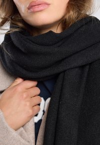 Zwarte gebreide sjaal om de nek gedrapeerd, met een zachte textuur. Gemaakt over een marineblauw shirt, met een deel van een beige jas zichtbaar.