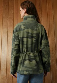 Kamouflagejacka i gröna nyanser, med hög krage, elastisk midja och stora ärmar, tillverkad av ett mjukt, strukturerat tyg.