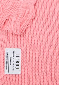 Il tessuto lavorato a maglia rosa presenta un motivo texturizzato con dettagli frangiati. È visibile un'etichetta con la scritta "LIL'BOO Danish Design Studios".
