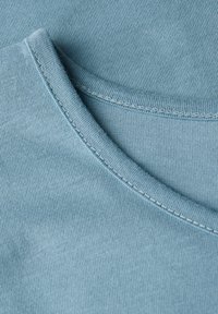 Detailní záběr na světle modrou bavlněnou látku s viditelným švem podél složených okrajů, zobrazující texturu a detaily materiálu trička.