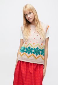 Mujer rubia con chaleco rosa de punto con patrón floral sobre una camiseta blanca y una falda roja, posando frente a un fondo liso.