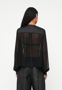 Blusa nera trasparente con scollatura rotonda e maniche lunghe a sbuffo. Presenta un giromanica nero opaco e un retro trasparente. Polsini con bottoni.