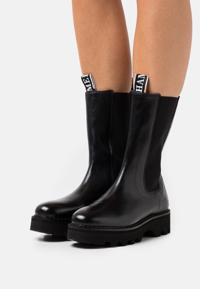 Black leather ankle boots with elastic side panels, rubber lug soles, and fabric pull tabs featuring contrasting white text. Smooth texture.