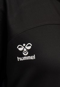 Black fabric with white Hummel logo featuring a stylized bee and brand name printed on the textile surface.