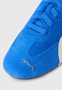 Blauwe suède sneakers met een gestructureerde bovenkant, platte veters, contrasterende beige accenten en een geborduurd logo nabij de neus.