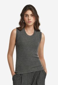 Ung kvinna med axellångt vågigt brunt hår som bär silvriga hoops örhängen och en åtsittande ärmlös ribbad grå topp med texturerade grå byxor.